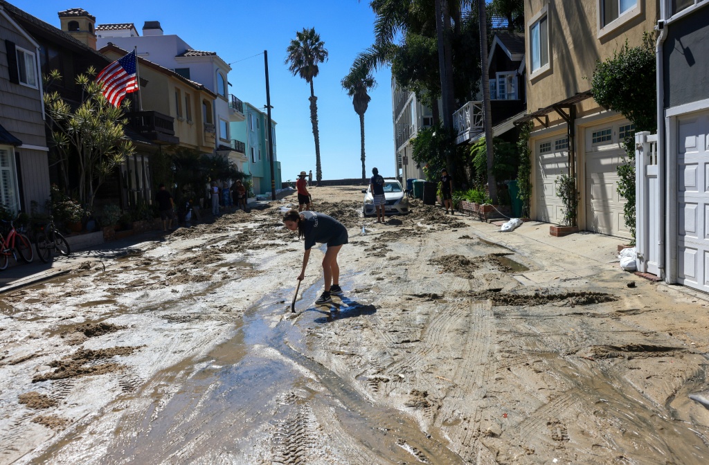 A woman shovels sand brought by the storm surge onto a street in Long Beach, California, which could see more coastal flooding into the weekend — David Swanson / AFP