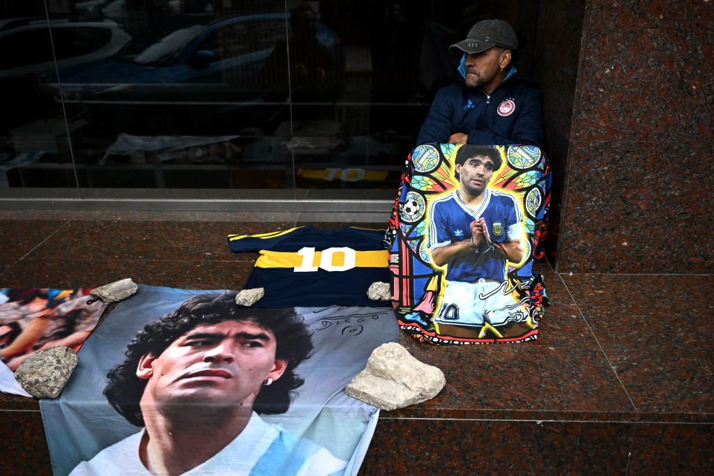 A supporter of Argentine football legend Diego Maradona sits beside portraits and memorabilia outside the courthouse during the final hearing of defense closing arguments in the trial over Maradona's 2020 death — Luis ROBAYO / AFP