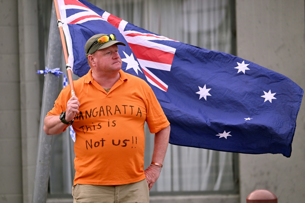 A man holds an Australian flag outside the office of Australian politician Helen Haines in the northern regional city of Wangaratta, about 250 kilometres north-east of Melbourne on October 20, 2026.  Australian independent lawmaker Helen Haines underwent surgery overnight but remains in a critical condition after being struck deliberately by a car in an attack, Prime Minister Anthony Albanese said on October 20. Political violence is rare in Australia, but law enforcement have documented a steady rise in th